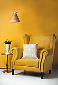 Yellow wing-back chair with pale scatter cushion, yellow tulips on side table and pendant lamp in front of yellow wall