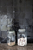 Seashells and sand in two jars in front of black wall tiles