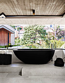 Dark, free-standing bathtub in the bathroom with concrete ceiling, concrete floor and garden view