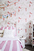 Bed with white and pink striped bed linen and wallpaper with bird motif in the girl's room