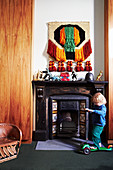 Toddler with scooter in front of an antique fireplace, toy dinosaurs and wall hanging