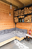 Sofa and side table on castors below firewood stacked in wall-mounted shelves in room with wooden walls