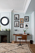 Teak desk next to marble fireplace, gallery of photographs and round mirror on mint-green wall