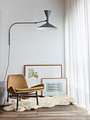 Designer chair on sheepskin rug, pictures leaning against wall and wall-mounted lamp next to window with floor-length curtains