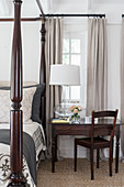 Table lamp on antique table and chair next to four-poster bed in country-house bedroom
