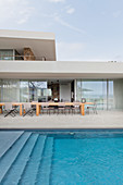 View across pool to long wooden table on stone-flagged terrace with glass sliding doors leading into house