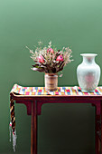 Console with table runner, vase and bouquet of flowers in front of green wall