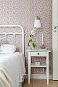 Flower in vase and brass candlestick on bedside table, crocheted blanket on metal bed in bedroom with floral wallpaper