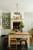 Soft green coloured kitchen with wooden stools and collection of plates stacked on open shelves