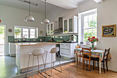 White kitchen with concrete floor and green splashback and narrow table with chairs on wooden floor in foreground