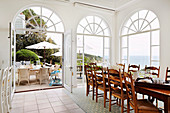 Long table with wooden chairs in front of arched window fronts with a view of the terrace and the sea