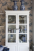 Blue-and-white crockery in classic display case against floral wallpaper
