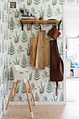 Chopping boards and leather apron hung from coat rack and baby's high chair in hallway with botanical wallpaper