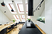 Island counter and dining table with black chairs in high-ceilinged room with skylights in sloping wall