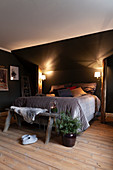 Box spring bed between rustic wooden columns below sloping ceiling lit by two wall-mounted lamps
