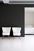 Two white retro armchairs against black wall with view into bedroom