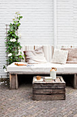 Rustic bench with seat cushion and scatter cushions against white-painted brick wall with old wooden crate used as table