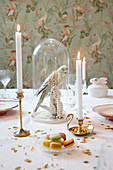 Vogelfigur mit Perlenkette unter Glashaube auf festlich gedecktem Tisch mit Kerzen, Macarons und  Goldsternen