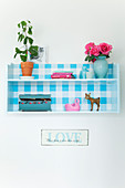Houseplant, a bouquet of roses, wooden boxes, and figures on a wall shelf with a blue and white checkered back wall