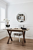 Books and orchid on wooden table, chair with striped upholstery and round mirror on wall