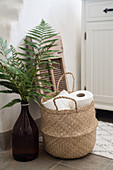 Fern leaves in large brown bottle and basket of toilet rolls in bathroom