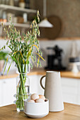 Fresh eggs in bowl, vase of grasses and jug on wooden table