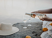 Woman pouring glasses of sparkling wine on rocks on lake shore