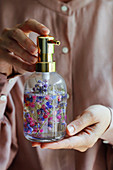 Hands holding soap dispenser of clear soap containing purple and pink dried flowers