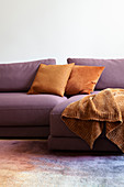 Orange blanket and scatter cushions on purple sofa on multicoloured rug