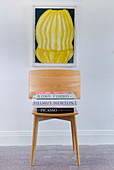 Books stacked on wooden chair below artwork on wall