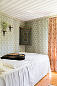 Bed with white cover and wall-mounted corner cabinet in bedroom with patterned wallpaper