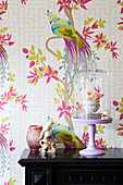 Rose in teacup under glass cover and bird ornament against wallpaper with bird motif
