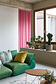 Grünes Sofa mit gelbem Kissen vor rosa Gardinen, neben Fenster mit Pflanzen