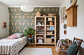 Single bed and cupboard with glass doors against patterned wallpaper in guest room