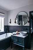 Washstand with marble top and countertop sink, round mirror and bathtub in bathroom with wainscoting