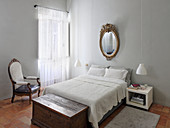 Gilt-framed mirror above double bed and trunk in bedroom with light grey walls
