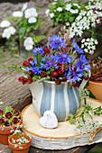 A small bouquet of cornflowers and crimson clovers in a pitcher on a disc of birch wood, a heart made of howlite crystal