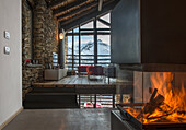 Kamin mit Feuer, im Hintergrund moderne Lounge in offenem Wohnraum mit Natursteinwänden und Holzbalkendecke