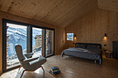 Polstersessel und Doppelbett vor Fensterfront im Schlafzimmer mit Holzverkleidung