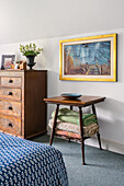 Painting above table with folded bedspreads and wooden chest of drawers