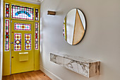 Floating shelf, round mirror and yellow front door with stained glass elements in hallway