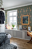 Radiator covers as shelf, above it painting with gold frame on wallpapered wall in a bedroom