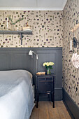 Bedside table next to a large bed in a bedroom with wainscoting and botanical wallpaper