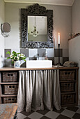Washstand with basket and linen curtain, above mirror with carved frame in a bathroom