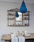 Rustikaler Holztisch mit Tischläufer und Geschirr, darüber blauer Kronleuchter und alter Bistroregal mit Spiegel