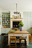 Soft green coloured kitchen with wooden stools and paltes stacked on open shelves