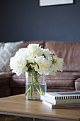 White peonies in a glass vase on a coffee table in front of a brown leather couch