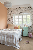 A bed and a mint green bedside table in a child's room with wallpaper and an apricot-coloured wall