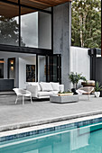 Outdoor furniture on terrace next to swimming pool outside two-storey house with glass walls