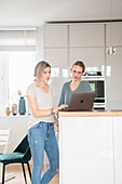 Two girlfriends are standing at the kitchen counter with laptop
